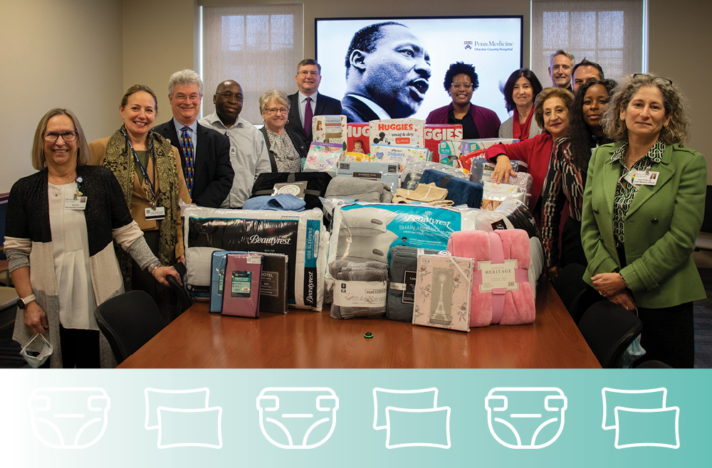 A group of people stands around a conference table covered with diaper boxes, blankets, and pillows. Behind them, a screen shows an image of Dr. Martin Luther King, Jr. 